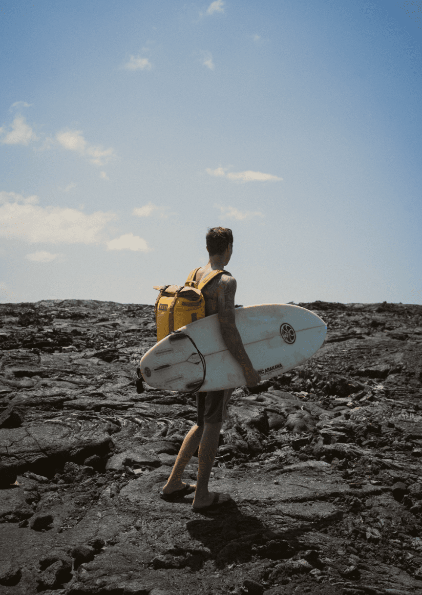 person carrying surfboard and backpack walking on dried lava